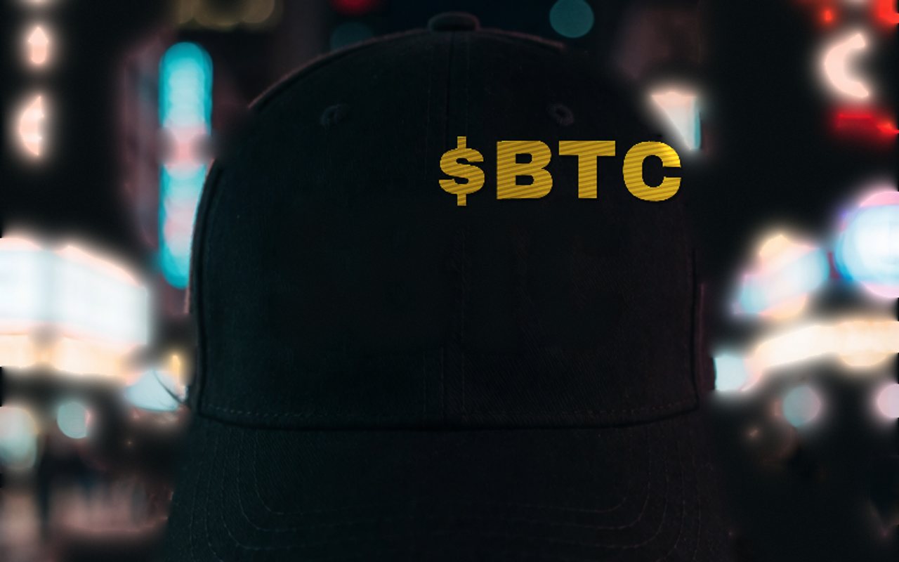 Person in low light wearing the black $BTC cap, orange embroidery catching the light